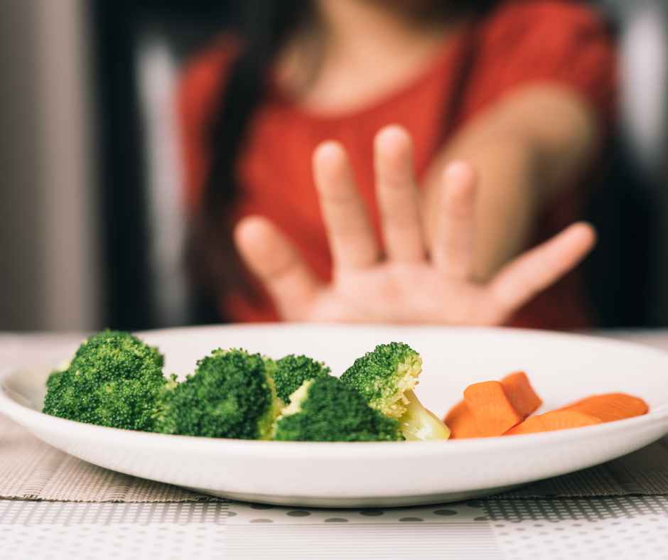 A smiling Picky Eater picking colorful vegetables off a plate shaped like a happy face, with a mom encouraging gently nearby.