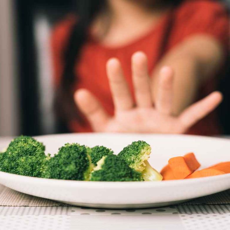 A smiling Picky Eater picking colorful vegetables off a plate shaped like a happy face, with a mom encouraging gently nearby.