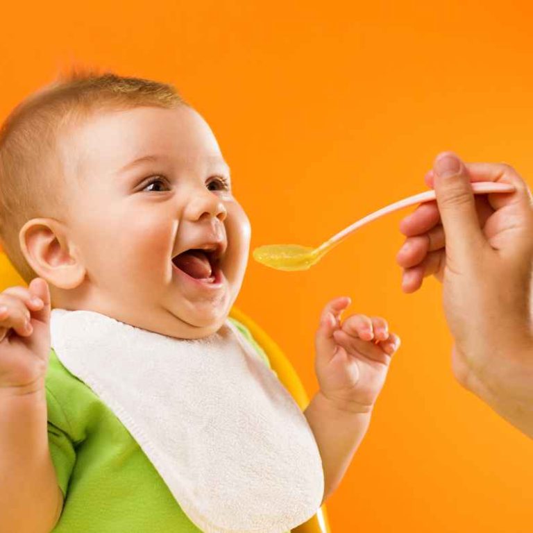 Starting solids for babies with mashed avocado served by mom using a spoon in a highchair